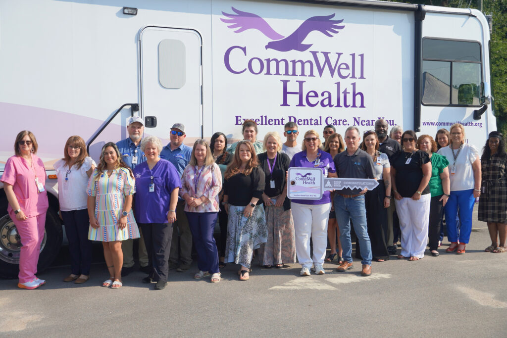 Group photo of approximately 25 CommWell Health staff members standing in front of a new white mobile dental health unit featuring the CommWell Health logo and the slogan "Excellent Dental Care. Near You." Team members smile toward the camera, while two individuals in the front hold a ceremonial oversized key recognizing the addition of the mobile unit, symbolizing expanded access to dental care across southeastern North Carolina.